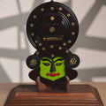 Decorative mask figure with green face and black hair on a wooden base against a neutral background